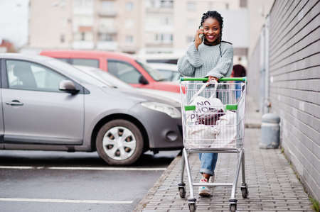African woman with shopping cart trolley posed outdoor market near car parking and speaking on phone.の写真素材