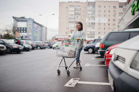 African woman with shopping cart trolley posed outdoor market near car parking.