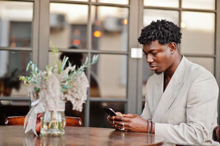 Stylish afro man in beige old school suit sitting on cafe with mobile phone. Fashionable young African male in casual jacket on bare torso.の写真素材