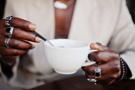 Stylish afro man in beige old school suit sitting on cafe with cup of coffee. Fashionable young...