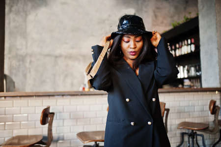 Charming african american woman model in black jacket, hat and waist bag relaxing in cafe during free time.の写真素材