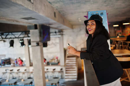 Charming african american woman model in black jacket relaxing in cafe during free time with mobile...