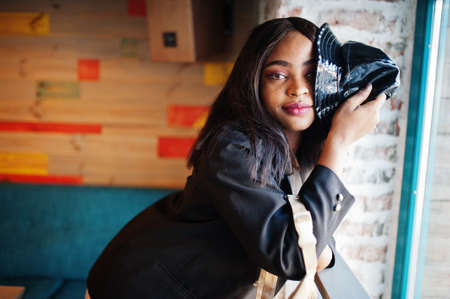 Charming african american woman model in black jacket and hat relaxing in cafe during free time.の写真素材