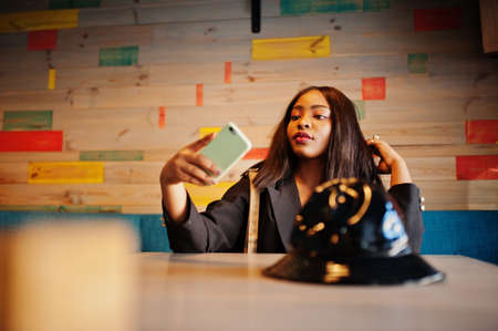 Charming african american woman model in black jacket relaxing in cafe during free time with mobile...