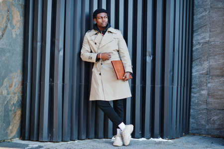 Handsome african american man posing outside in black hat and beige coat with folder in hand.