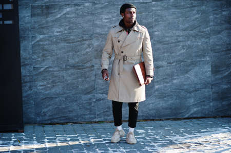 Handsome african american man posing outside in black hat and beige coat with folder in hand.の写真素材