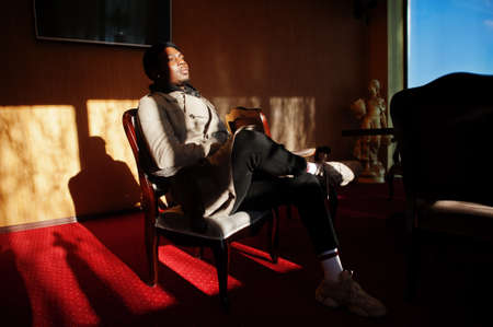 Handsome african american man posing inside room with sunlight shadows in black hat and beige coat.