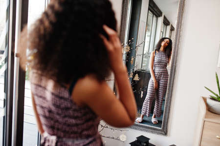 Charming elegant young african american woman with long curly hair wearing jumpsuit looking at mirror.の写真素材