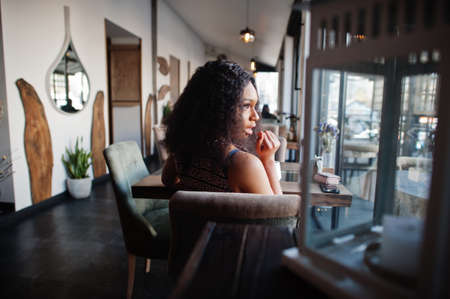 Ð¡harming elegant young african american woman with long curly hair wearing jumpsuit sitting at cafe indoor.の写真素材