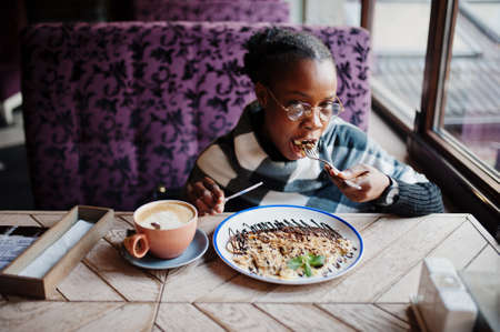 African woman in checkered cape and eyeglasses sitting at cafe and eating dessert.の写真素材