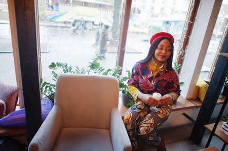 Enthusiastic african american woman in trendy colored outfit with red beret chilling in cozy cafe...