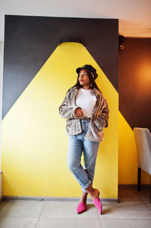Fashionable african american woman in black cap chilling in cozy cafe and fur against yellow wall.