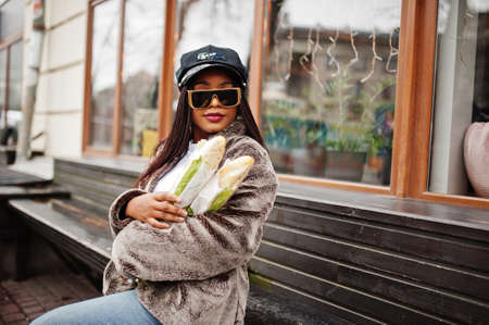 Fashionable african american woman in black cap, sunglasses and fur with two baguette rolls at...