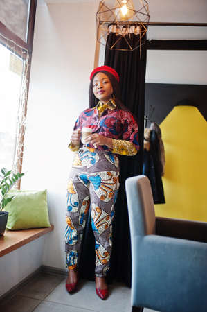 Enthusiastic african american woman in trendy colored outfit with red beret chilling in cozy cafe with cup of hot latte in hands.の写真素材