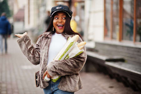 Fashionable african american woman in black cap and fur with two baguette rolls at hands.
