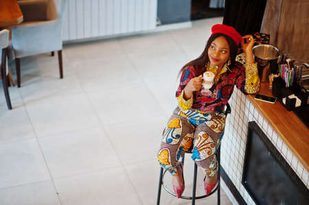 Enthusiastic african american woman in trendy colored outfit with red beret chilling in cozy cafe, sitting on bar counter.の写真素材