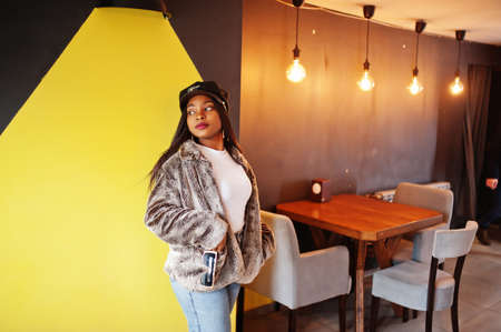 Fashionable african american woman in black cap chilling in cozy cafe and fur against yellow wall.