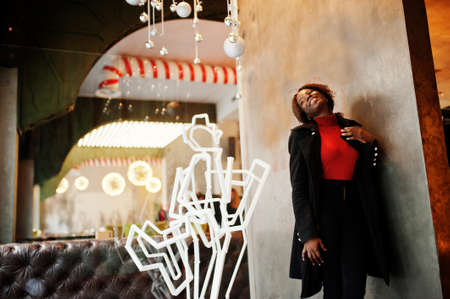Portrait of a curly haired african woman wearing fashionable black coat and red turtleneck posing at cafe.の写真素材