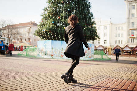 Portrait of a curly haired african woman wearing fashionable black coat and red turtleneck posing outdoor against main new year tree of city.の写真素材