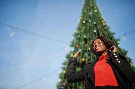 Portrait of a curly haired african woman wearing fashionable black coat and red turtleneck posing outdoor against main new year tree of city.の写真素材