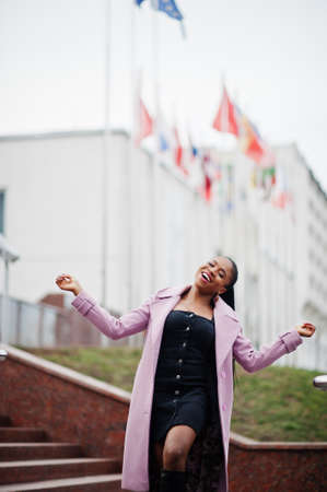 Young stylish beautiful african american woman in street, wearing fashion outfit coat, against flags...