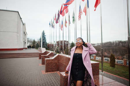 Young stylish beautiful african american woman in street, wearing fashion outfit coat and eyeglasses, against flags of different countries of the world.の写真素材