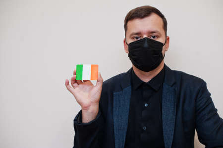 European man wear black formal and protect face mask, hold Ireland flag card isolated on white...