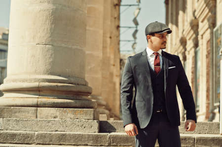 Portrait of retro 1920s english arabian business man wearing dark suit, tie and flat cap near old columns.の写真素材