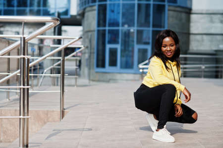 Hipster african american girl wearing yellow hoodie posing at street against office building with blue windows.の写真素材