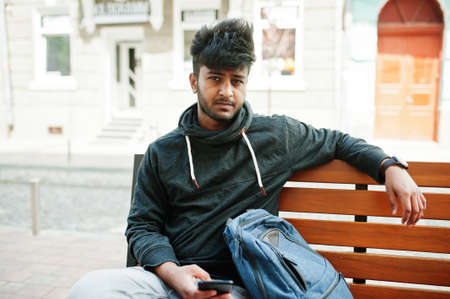 Portrait of young stylish indian man model pose in street, sitting on bench and hold smartphone at...
