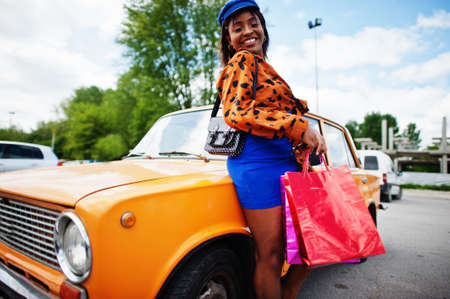 Beautiful african american lady with shopping bags standing near orange classic retro car.