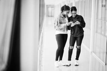 Two african woman friends in jeans jacket using mobile phones indoor together.の写真素材