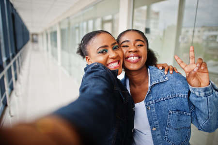 Two african woman friends in jeans jacket making selfie by camera indoor together.の写真素材