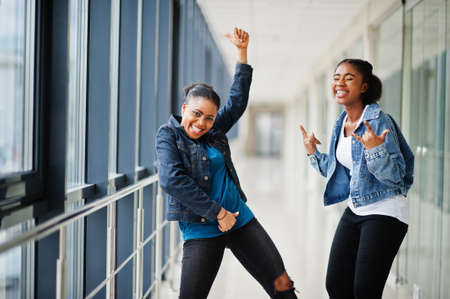 Two african woman friends in jeans jacket show rock fingers indoor together.の写真素材
