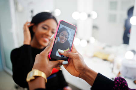 African American woman applying make-up by make-up artist at beauty saloon. Artist make photo on mobile phone of her work.の写真素材