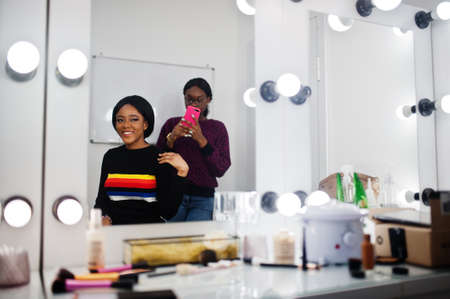 African American woman applying make-up by make-up artist at beauty saloon. Artist make photo on mobile phone of her work.の写真素材