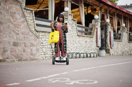 Beautiful african american woman using hoverboard and mobile phone. Black girl with yellow cloth eco...