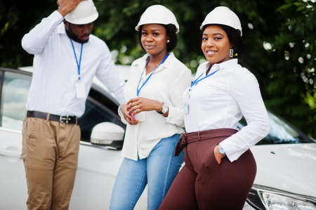 African american technician in white helmets near car. Group of three black engineers meeting.