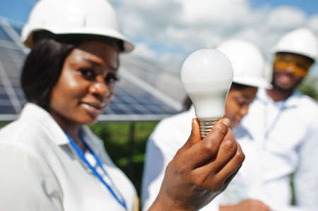 African american technician checks the maintenance of the solar panels. Group of three black engineers meeting at solar station. Hold led bulb.の写真素材