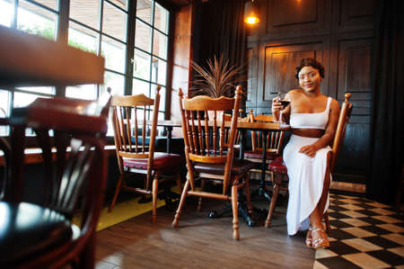 African american woman, retro hairstyle in white dress at restaurant with glass of wine.