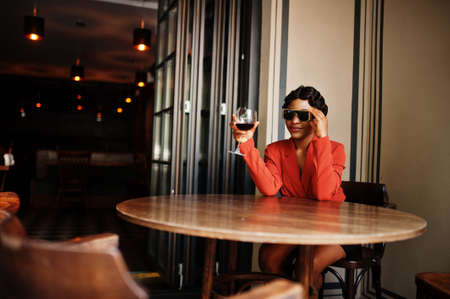 Portrait of african american woman, retro hairstyle with eyeglasses, wear orange jacket posing at restaurant with glass of wine.の写真素材