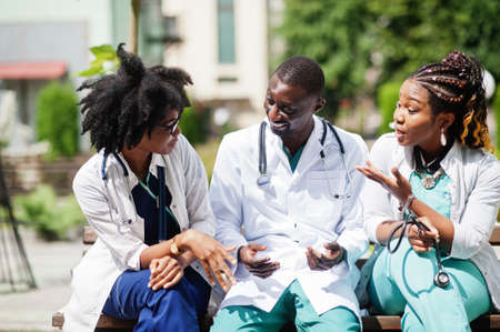 Three African American group doctors with stethoscope wearing lab coat sitting on bench.の写真素材