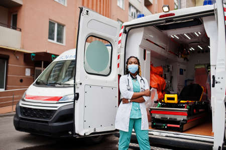 African american female paramedic in face protective medical mask standing in front of ambulance car.の写真素材