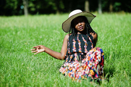 Portrait of gorgeous african american woman 20s in summer hat sitting at green grass in park.の写真素材