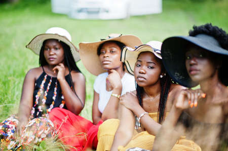 Group of four gorgeous african american womans wear summer hat sitting at green grass in park.の写真素材