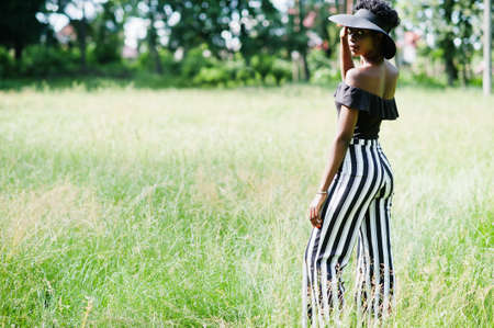 Portrait of gorgeous african american woman 20s in wear in black and white stripe pants and summer hat posing at green grass in park.の写真素材