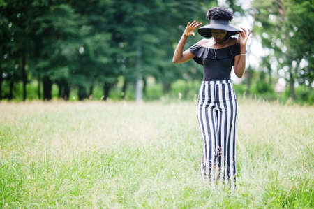 Portrait of gorgeous african american woman 20s in wear in black and white stripe pants and summer hat posing at green grass in park.の写真素材