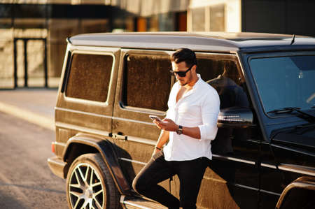 Solid asian man in white shirt and sunglasses posed near black mafia suv car with mobile phone.