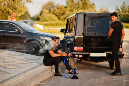 Two asian brothers man wear on all black posed near suv cars and smoke hookah.