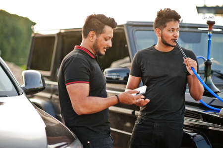 Two asian brothers man wear on all black posed near suv cars and smoke hookah.